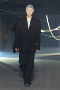 Model walks the runway in an oversized black suit with a white shirt; sheer hood covers the head.