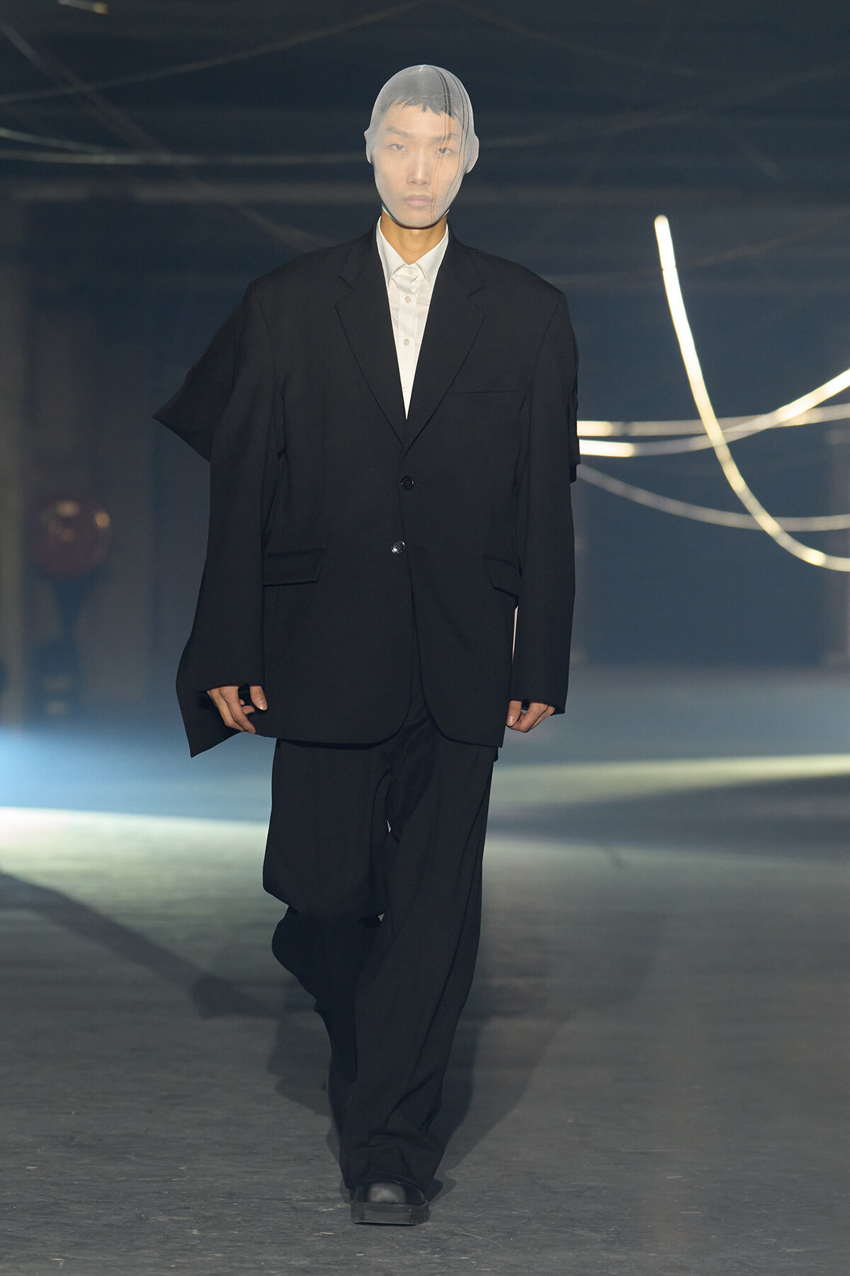 Model walks the runway in an oversized black suit with a white shirt; sheer hood covers the head.