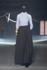 Model on a runway wearing a light blue button-down shirt tucked into a long, voluminous black skirt; face obscured by a dark mask/hood.