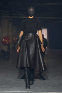 Model walking a dimly lit runway in an all-black ensemble: fitted black top, voluminous black leather skirt, knee-high black boots, and a dark mask covering the face.