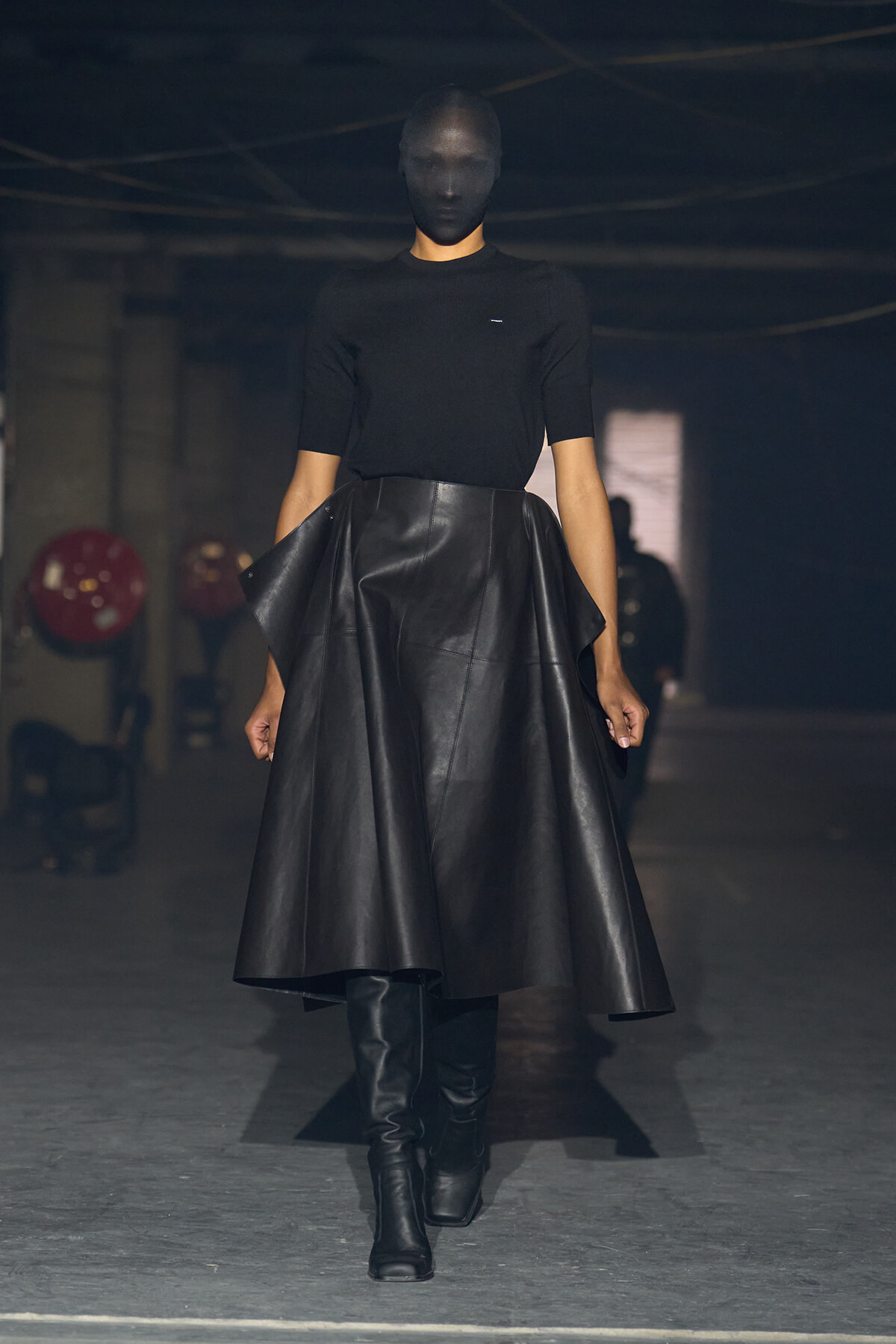 Model walking a dimly lit runway in an all-black ensemble: fitted black top, voluminous black leather skirt, knee-high black boots, and a dark mask covering the face.