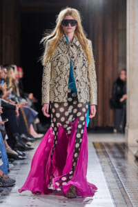 Model on runway wearing snake-print trench over blue top, paired with sheer black polka-dot skirt and bright pink cape-like hem; oversized sunglasses.