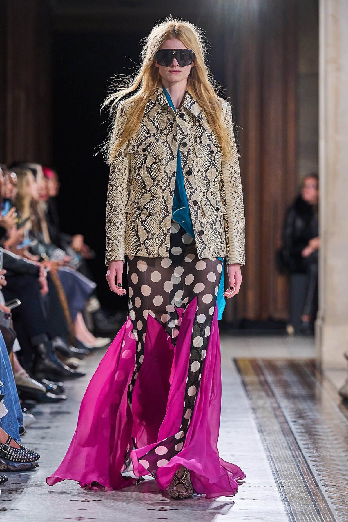 Model on runway wearing snake-print trench over blue top, paired with sheer black polka-dot skirt and bright pink cape-like hem; oversized sunglasses.