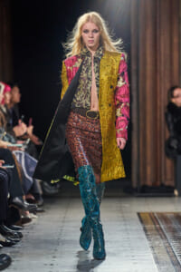 Model walks a fashion runway wearing a boldly patterned gold, pink, and black coat over a sheer patterned blouse and sequined skirt, teal snakeskin boots visible.