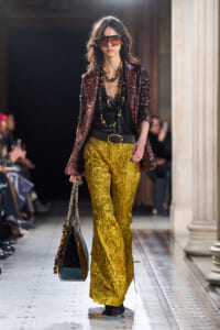 Model walking a runway in gold brocade trousers, a black top, a burgundy sequin blazer, large beaded necklaces, oversized sunglasses, and a patterned tote.