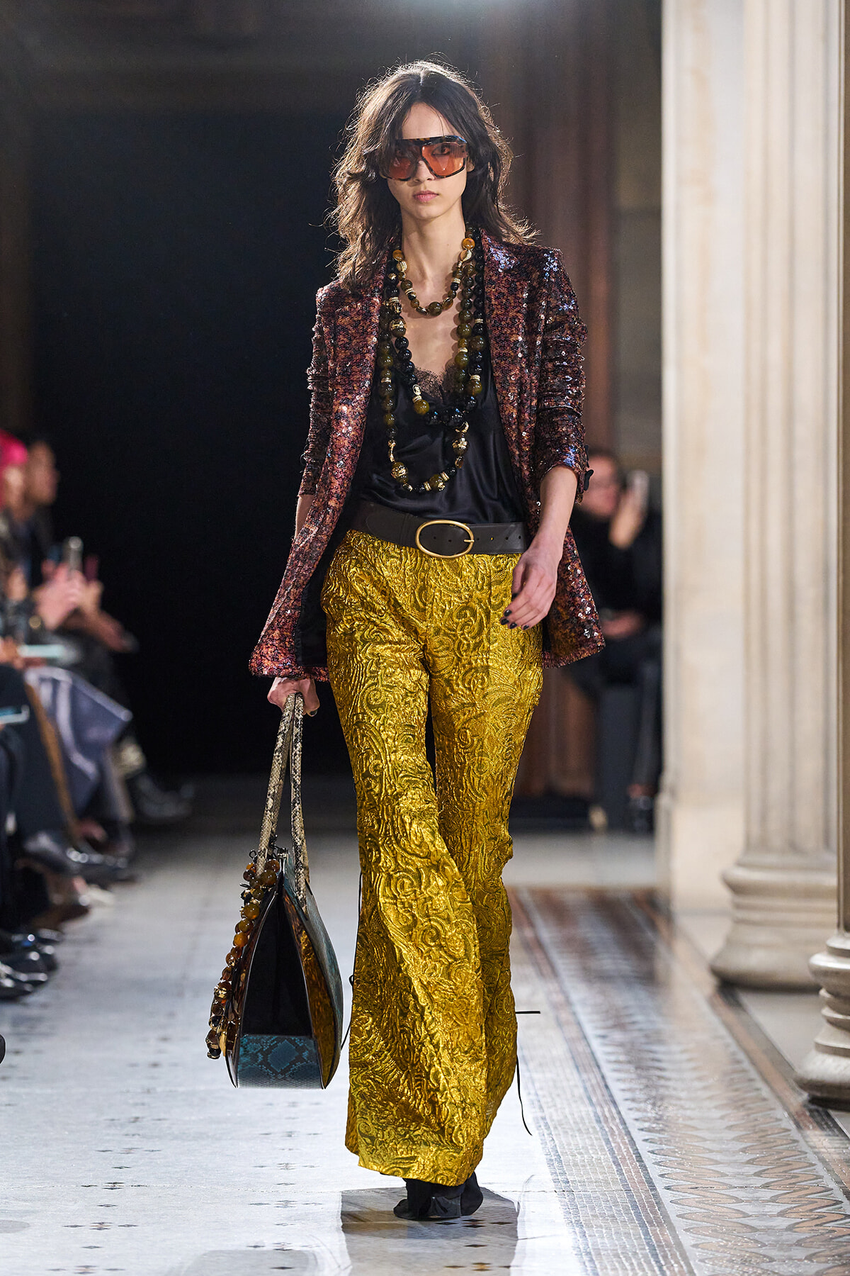 Model walking a runway in gold brocade trousers, a black top, a burgundy sequin blazer, large beaded necklaces, oversized sunglasses, and a patterned tote.