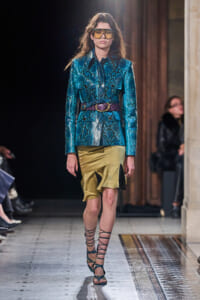 Model on a runway wearing a metallic blue snakeskin blazer with a purple belt, a gold satin skirt with black panels, oversized yellow goggles, and strappy gladiator sandals.