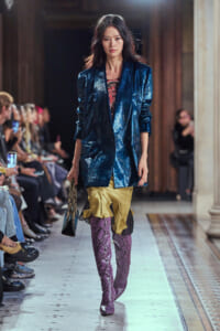 Female model walks a runway in a metallic blue blazer over a gold ruffled skirt, purple snakeskin boots, and a snakeskin bag.