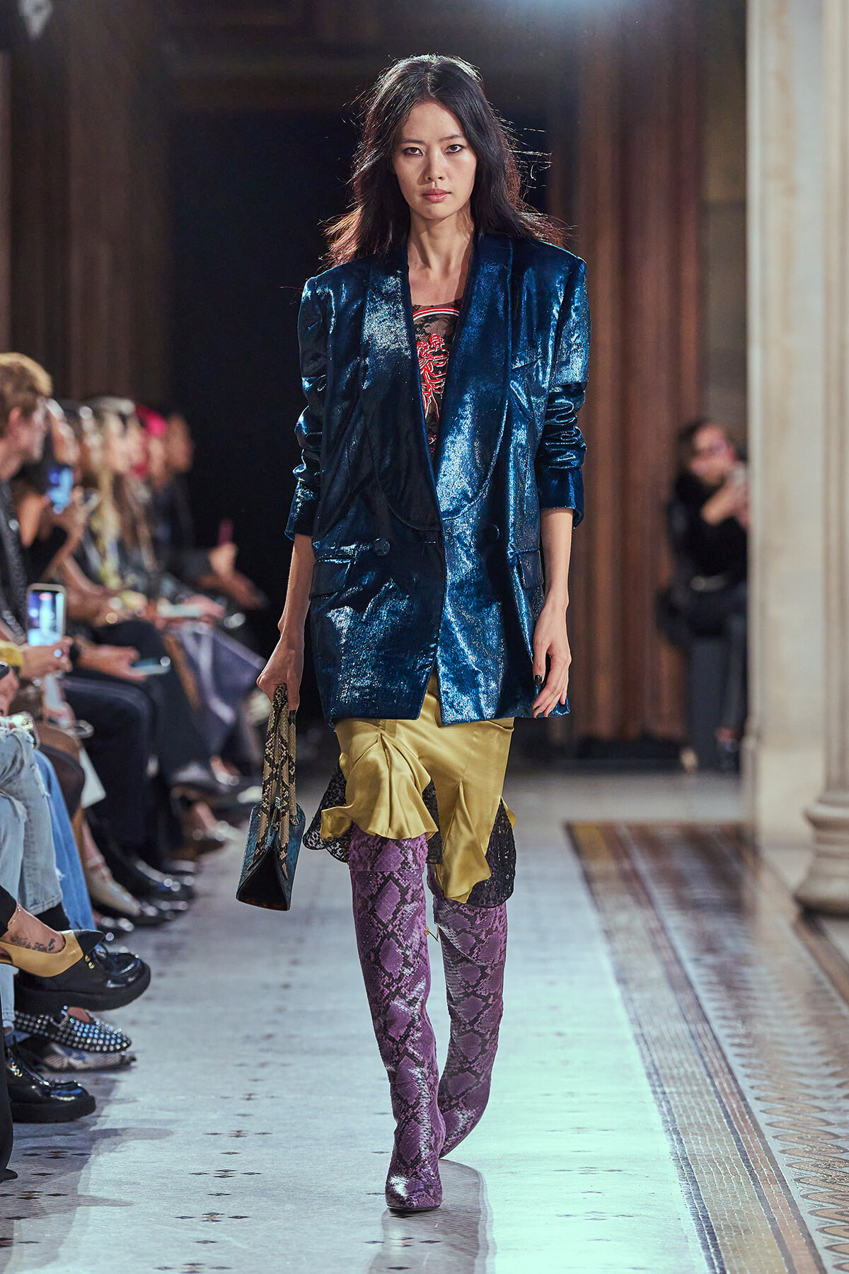 Female model walks a runway in a metallic blue blazer over a gold ruffled skirt, purple snakeskin boots, and a snakeskin bag.