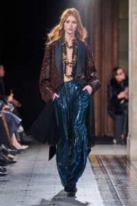 Model walking a runway in a burgundy sequined coat with a black satin lapel and large beaded necklaces over teal metallic pants.