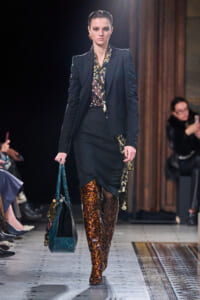 Model walks a runway in a black blazer, printed blouse, and black pencil skirt with a snakeskin knee-high boot duo; teal handbag in hand.