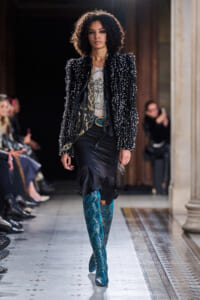 Model on runway wearing a sequined black jacket, a graphic tee, a fitted black skirt, and turquoise snakeskin knee-high boots.