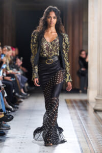 Model walking a fashion runway in a sheer black polka-dot maxi skirt over pants, paired with a gold-snake print top and black blazer with a double-ring belt.