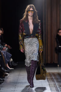 Model walks a runway in a deep-plunging metallic jacket, leopard-print skirt, and purple snakeskin boots, carrying a patterned bag and wearing large sunglasses.