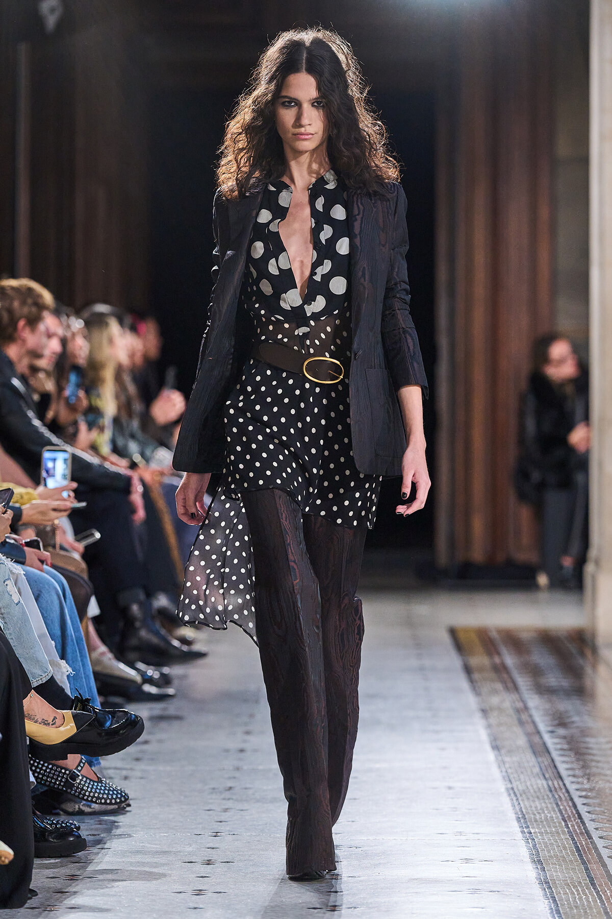 Model walking the runway in a black polka-dot dress with a deep neckline, paired with a dark blazer and a tan belt around the waist.