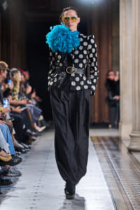 Model walking a runway in a black polka-dot blazer with a large blue flower, gold belt, and oversized yellow sunglasses, paired with wide-leg pinstripe pants. Audience visible in background.]]}{