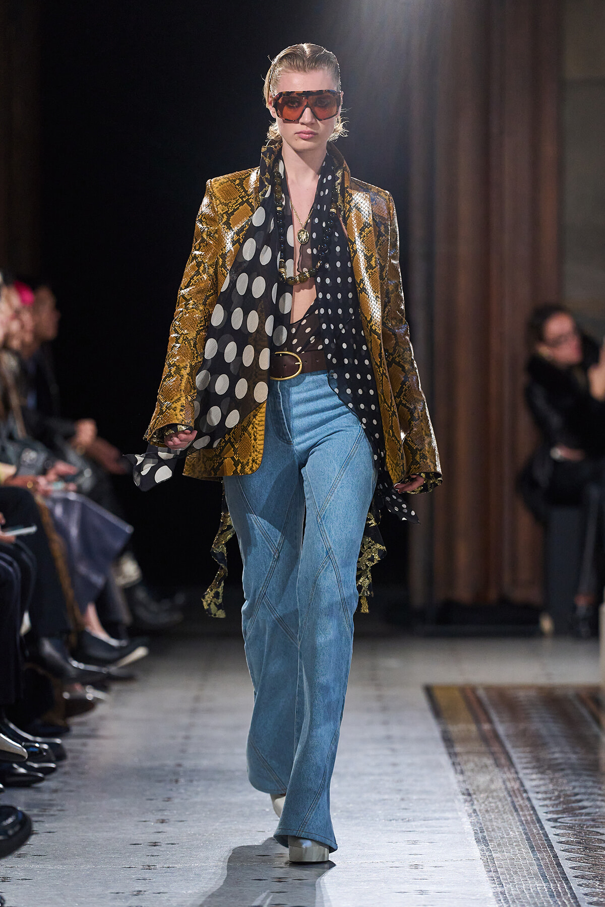 Model walking a runway in a metallic snakeskin blazer, large polka-dot scarf, blue bootcut jeans, and oversized sunglasses.