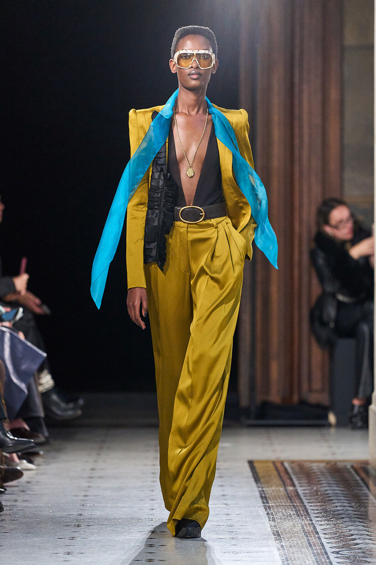 Model on runway wearing a mustard-yellow suit with a deep V black top, blue scarf, large belt, and gold accessories; sunglasses add retro flair.