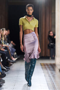 Model walking a runway in a lime cropped polo shirt, lilac satin skirt with a ruffled hem, and teal snakeskin boots.