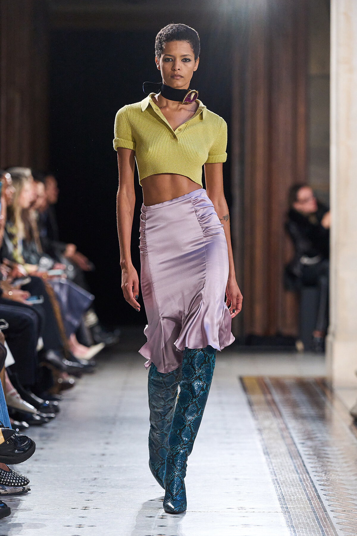 Model walking a runway in a lime cropped polo shirt, lilac satin skirt with a ruffled hem, and teal snakeskin boots.