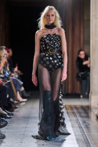 Model walking a runway in a black strapless sheer gown with a large polka-dot pattern and a brown belt.
