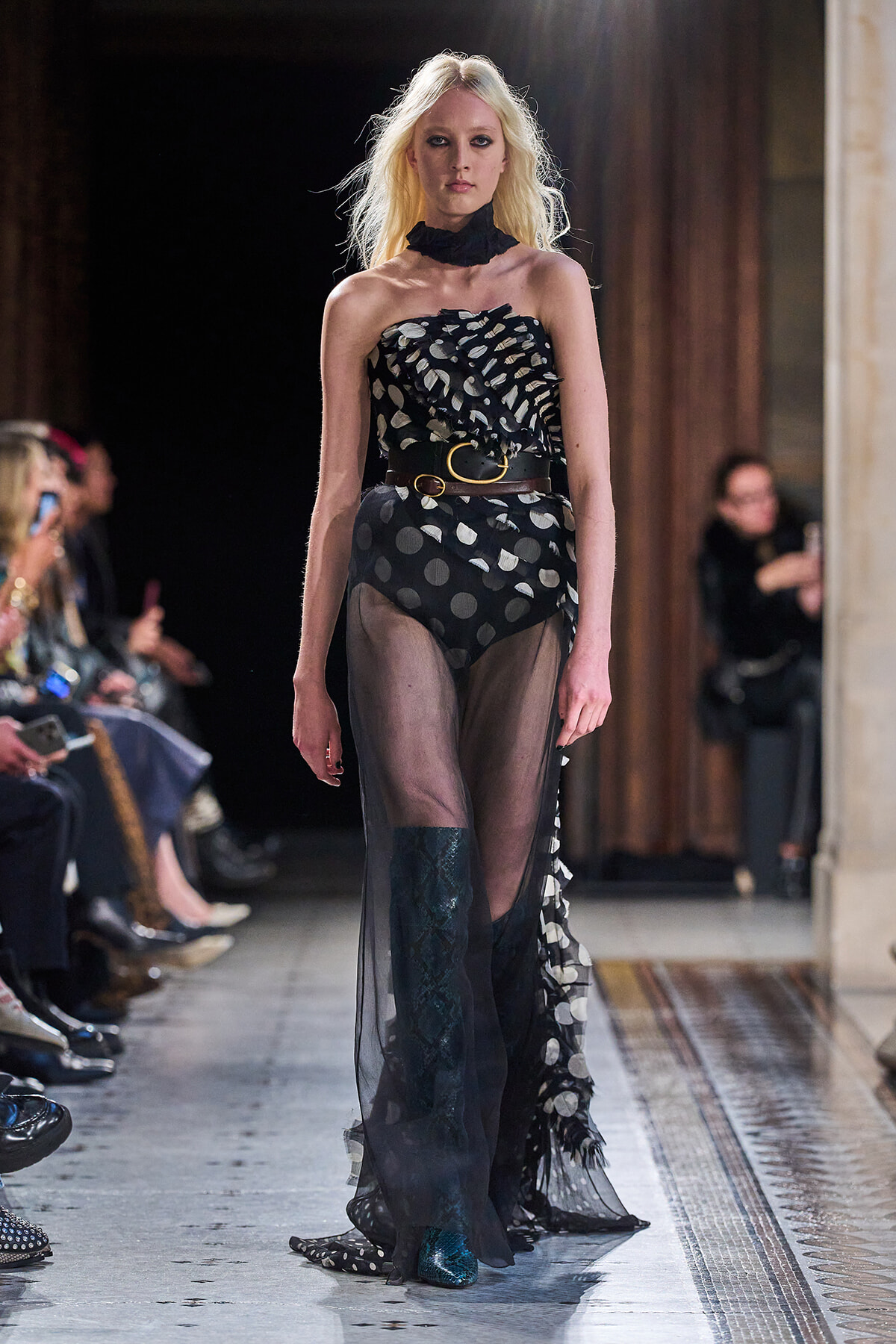 Model walking a runway in a black strapless sheer gown with a large polka-dot pattern and a brown belt.