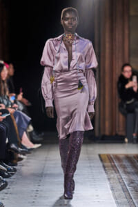 Model walks a lavender satin dress with puff sleeves on a runway, pairing it with knee-high snakeskin boots.
