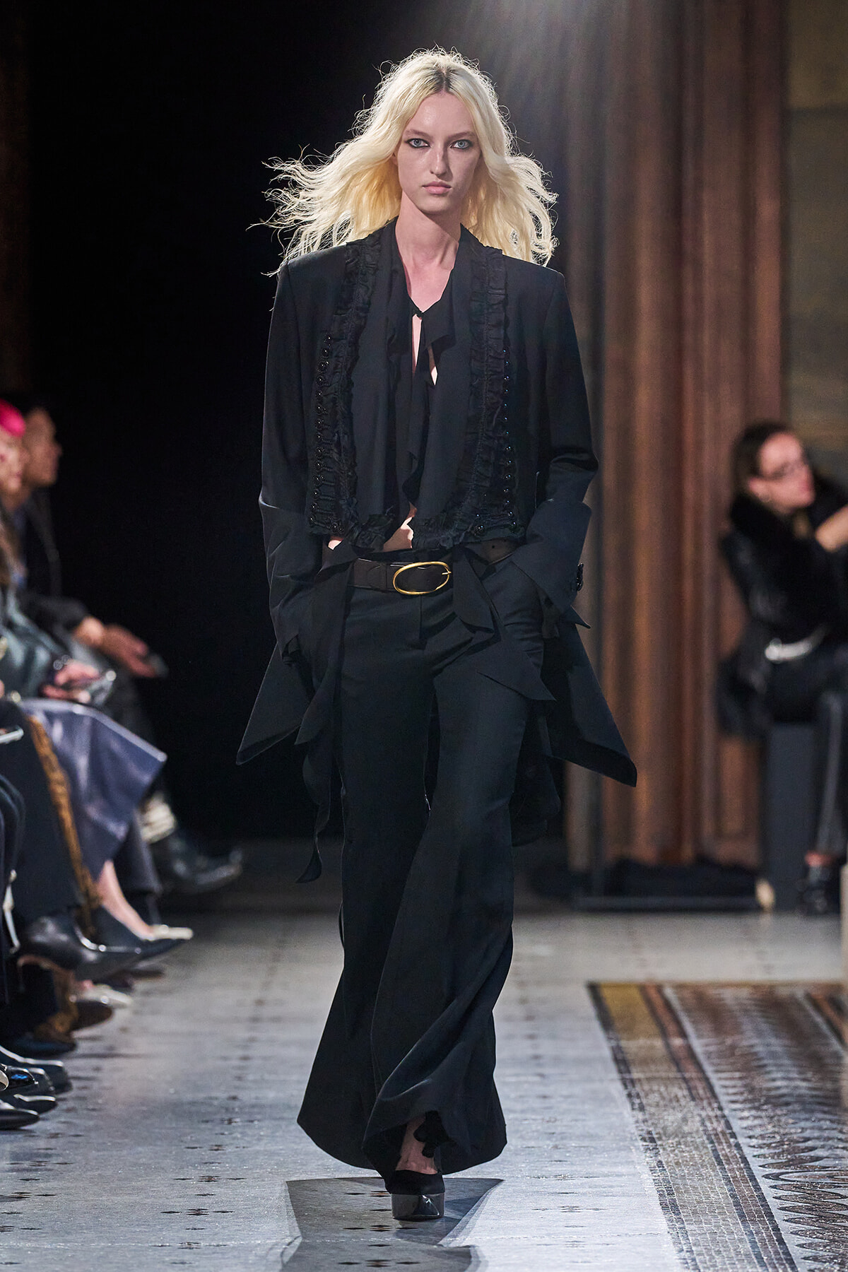 Model walks a runway in a black tailored suit with ruffled vest and belt, foregrounded by dramatic lighting and an audience on either side.