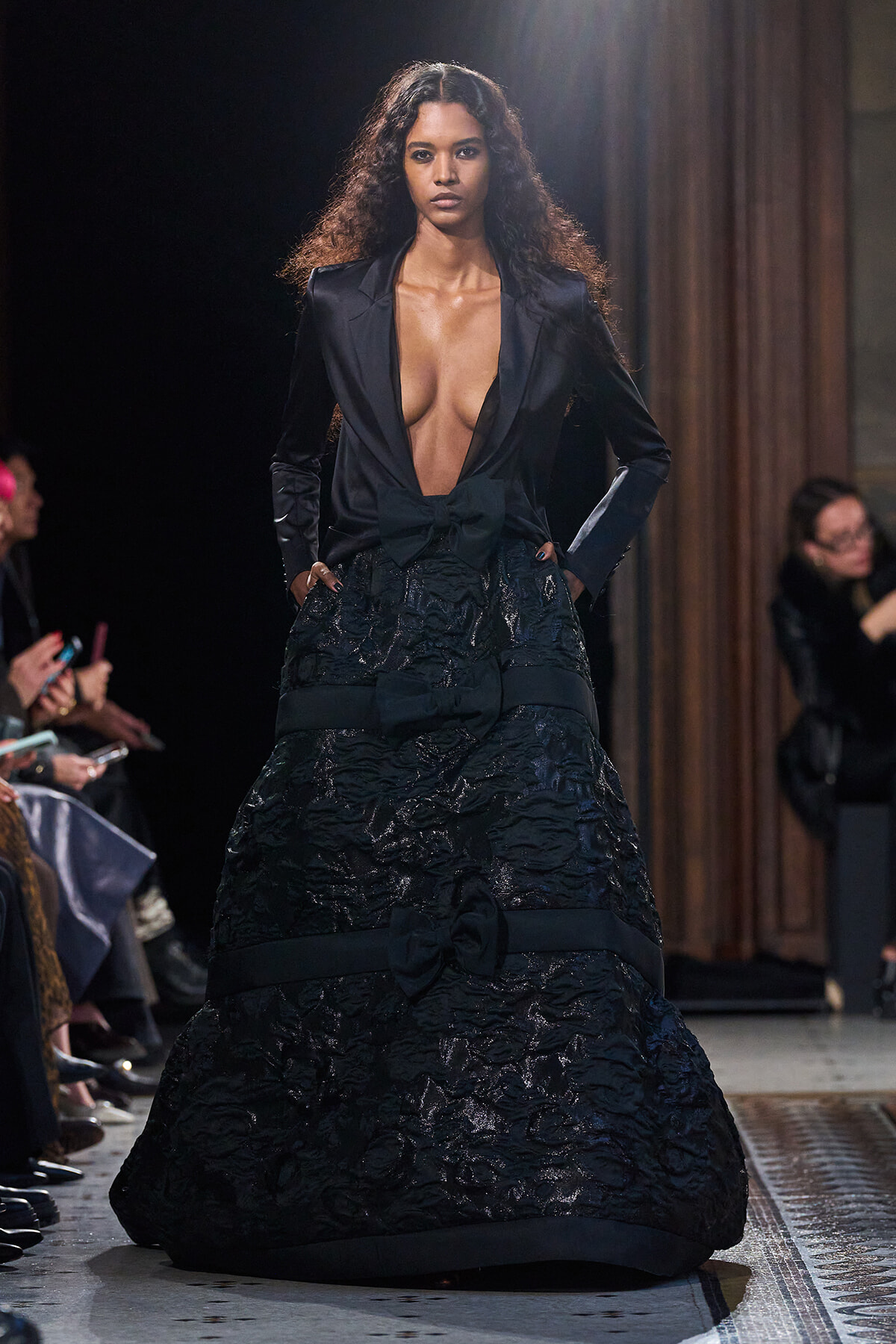 Model on a runway wearing a black satin blazer with a deep plunging neckline and a full, textured black skirt with bow accents.
