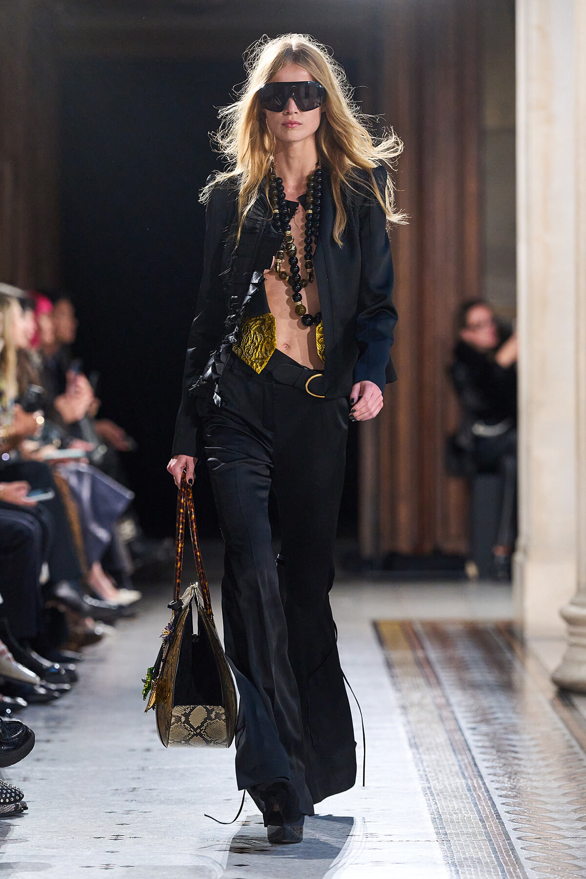 Runway model in a black suit with gold accents, oversized sunglasses, and long beaded necklaces, carrying a snakeskin tote.