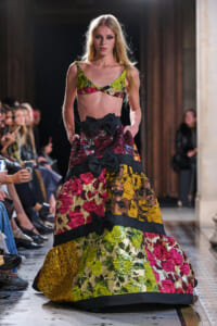 Model walks a runway wearing a vibrant patchwork floral ball gown with bold black bows and a matching bralette top under bright lighting.