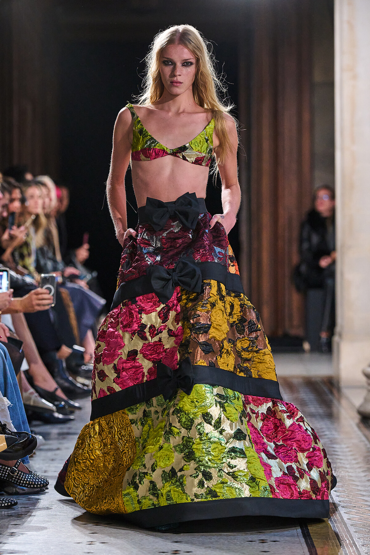 Model walks a runway wearing a vibrant patchwork floral ball gown with bold black bows and a matching bralette top under bright lighting.