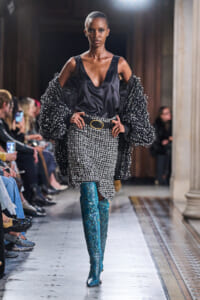 Model walks a runway in a black satin top, oversized beaded jacket, and a tweed-like skirt with a gold-buckled belt; teal snakeskin boots dominate the look.