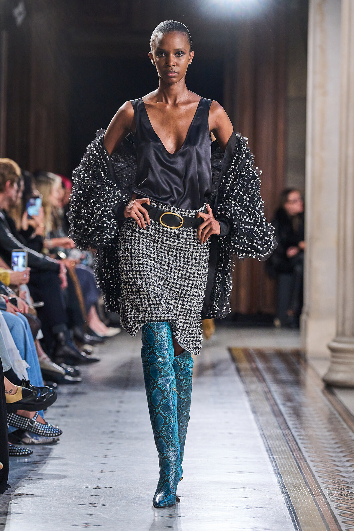 Model walks a runway in a black satin top, oversized beaded jacket, and a tweed-like skirt with a gold-buckled belt; teal snakeskin boots dominate the look.