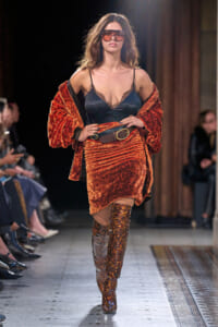 Model walks runway in orange velvet suit with black lace camisole and oversized brown belt, paired with leopard-print knee-high boots and red sunglasses.