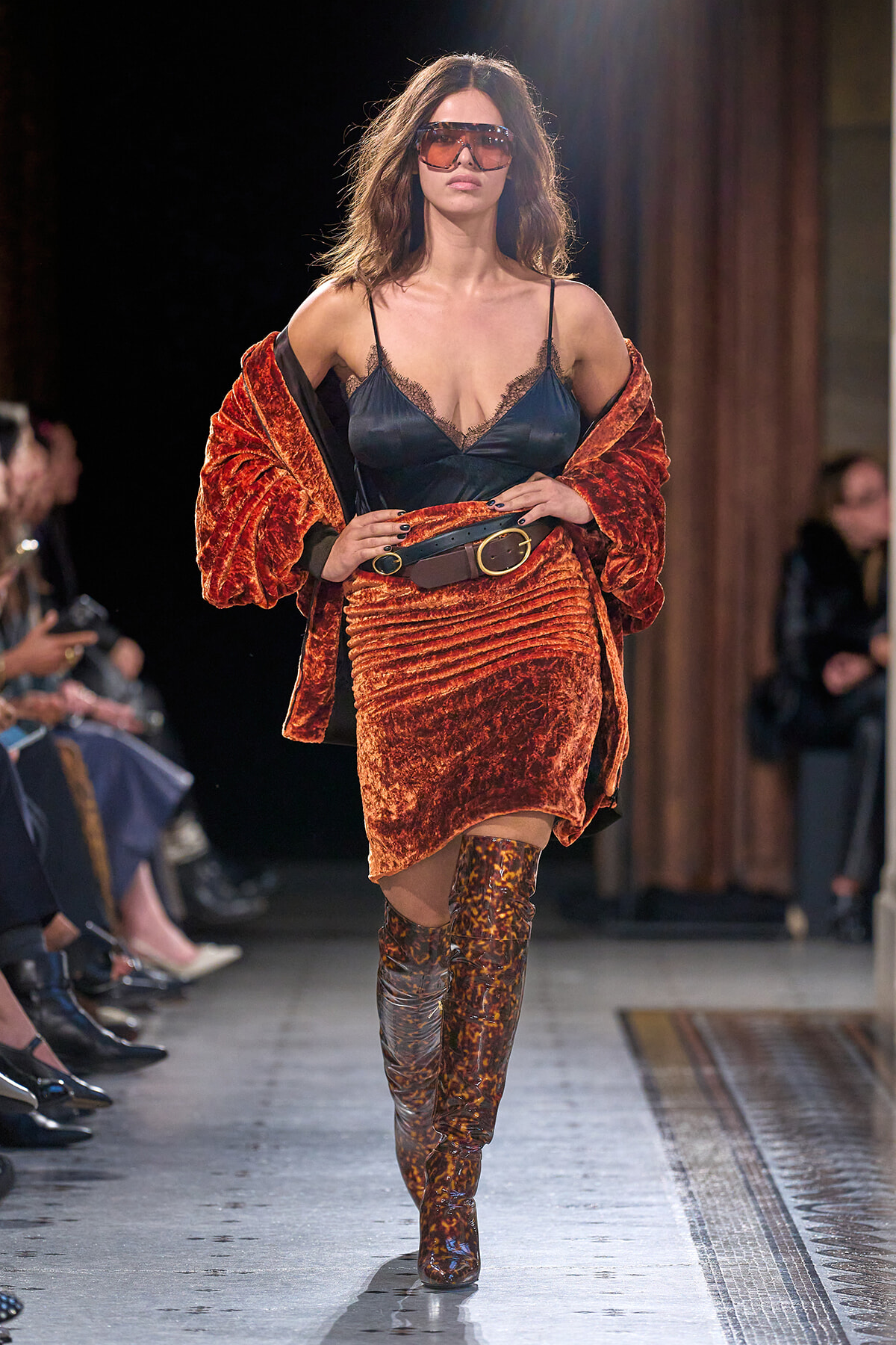 Model walks runway in orange velvet suit with black lace camisole and oversized brown belt, paired with leopard-print knee-high boots and red sunglasses.