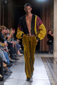 Male model walks runway in gold velvet pants, black plunging shirt, and a patterned yellow burgundy jacket with a brown belt and gold chain necklace.