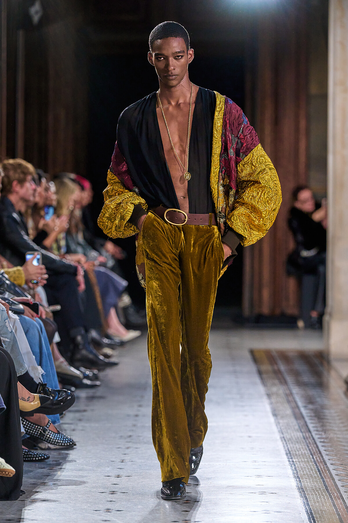 Male model walks runway in gold velvet pants, black plunging shirt, and a patterned yellow burgundy jacket with a brown belt and gold chain necklace.