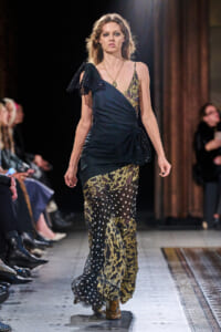 Runway model walking in a dark, layered evening gown with gold lace appliqué and a sheer polka-dot overlay at the hem, one-shoulder bow detail visible on the left shoulder.