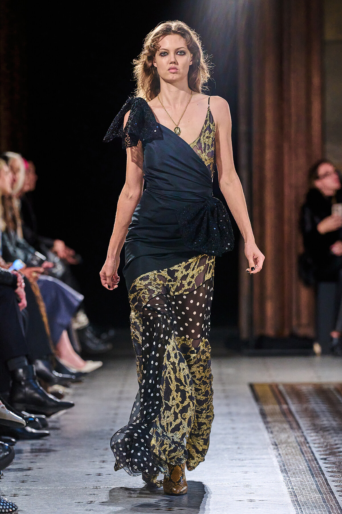 Runway model walking in a dark, layered evening gown with gold lace appliqué and a sheer polka-dot overlay at the hem, one-shoulder bow detail visible on the left shoulder.