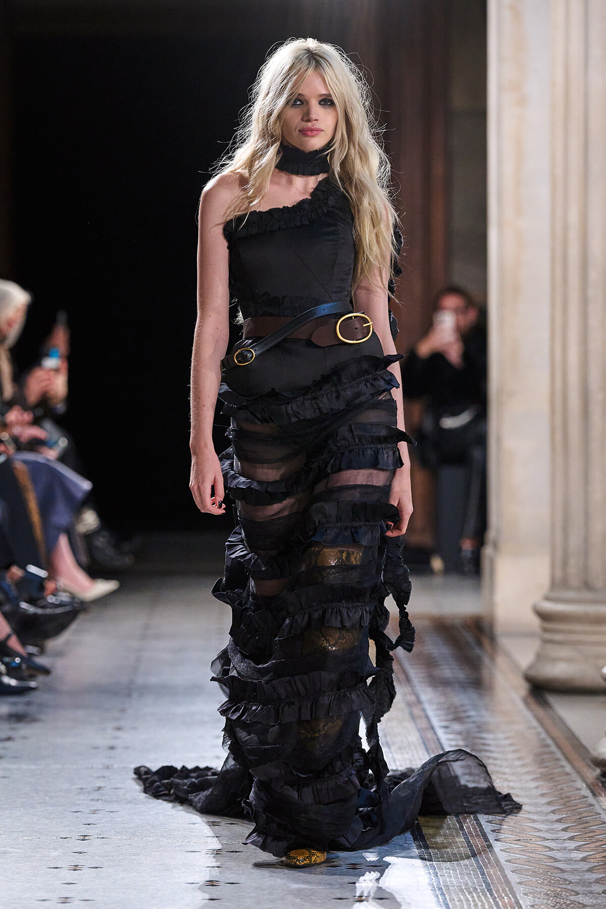 Model walking the runway in a black, multi-layered ruffled gown with a wide brown belt and gold buckle.