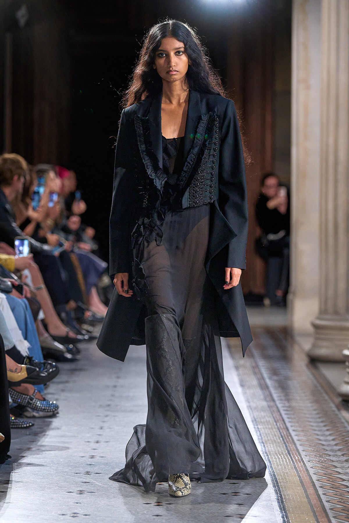 Model walks a runway in an all-black ensemble: structured coat with ruffled corset-like detailing and sheer, flowing pants, snakeskin boots visible at the hem.