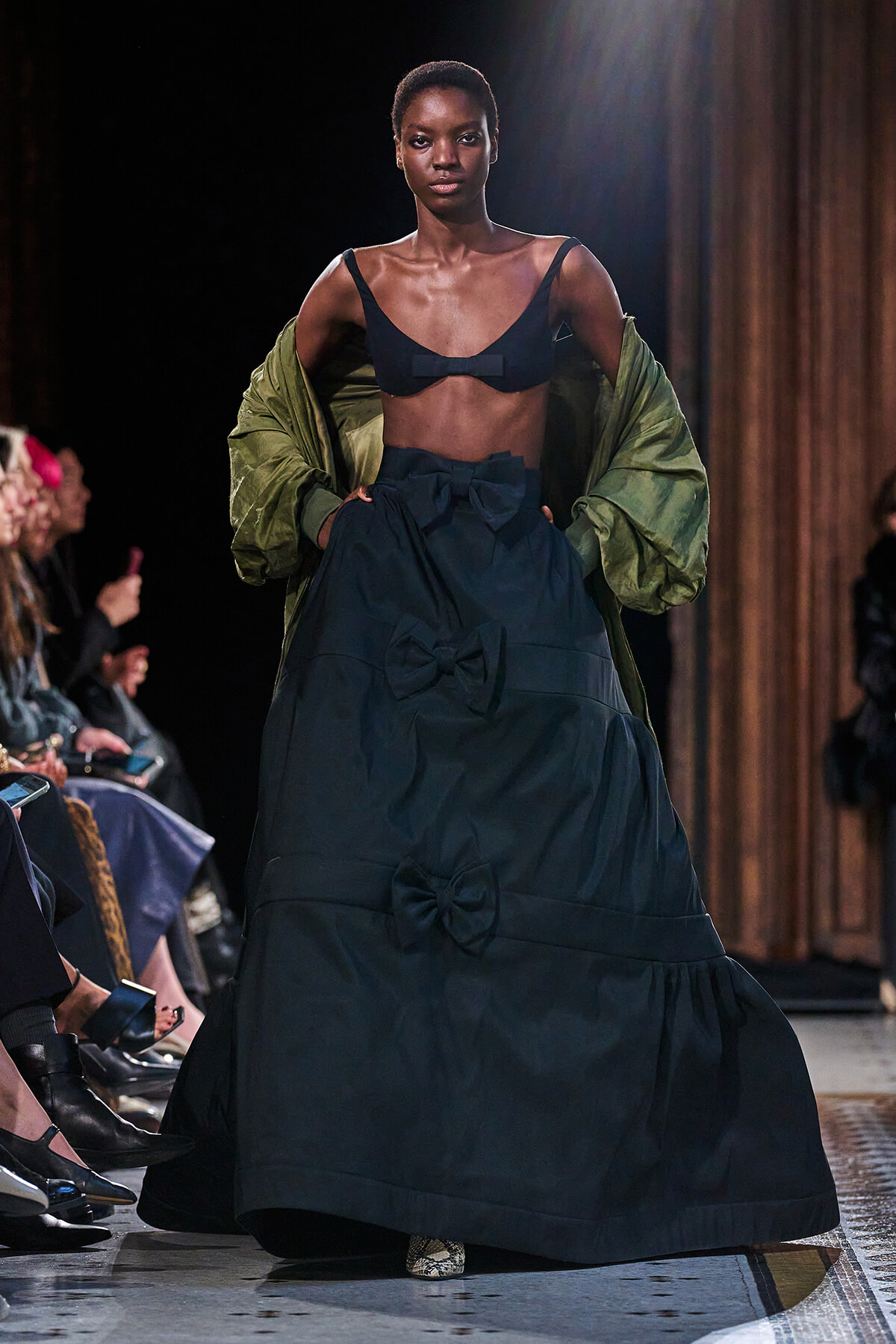 Model walks the runway in a navy, bow-accented gown with a cropped navy top, olive-green jacket draped over shoulders, audience along the catwalk.