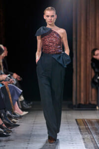 Model walks a runway in a one-shoulder sequined red top with dark high-waisted trousers and a decorative bow at the waist.