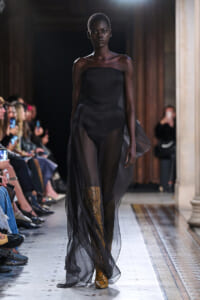 Black strapless dress with sheer overlay walking a fashion runway; gold snakeskin boots visible; audience on both sides watching.