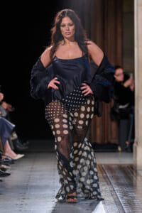 Plus-size model walking a runway in a navy satin slip dress with a sheer polka-dot overlay skirt and a dark bolero jacket.