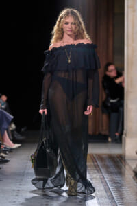 Model walking a runway in a sheer black off-shoulder gown with a ruffled neckline, gold pendant necklace, and snakeskin boots, carrying a black handbag.