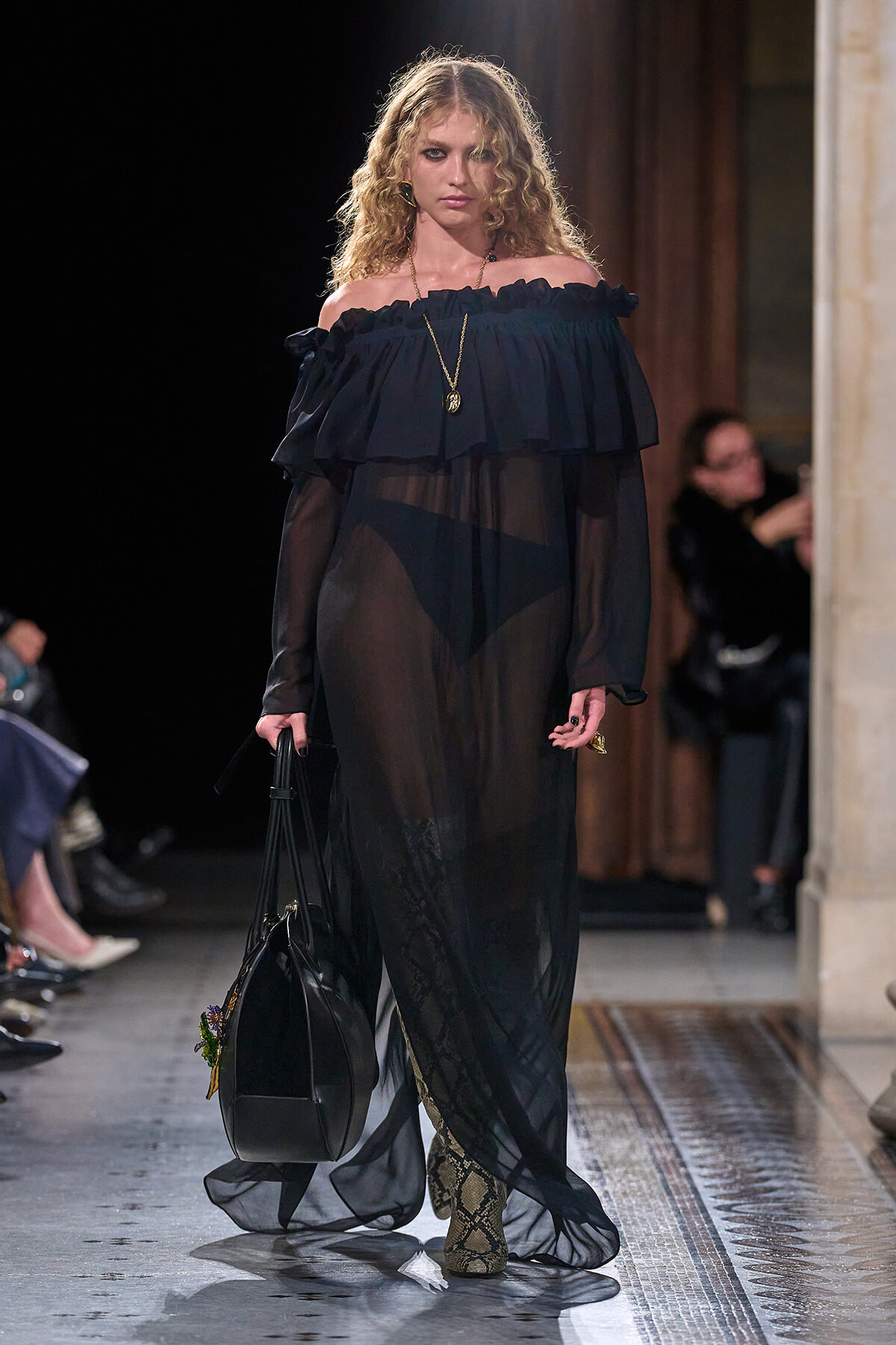 Model walking a runway in a sheer black off-shoulder gown with a ruffled neckline, gold pendant necklace, and snakeskin boots, carrying a black handbag.