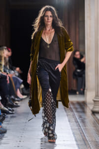 Model walking the runway in an olive-brown velvet coat over a sheer black top and polka-dot sheer pants with a leaf pattern detail.