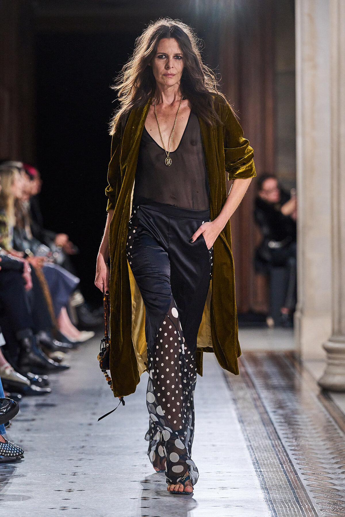 Model walking the runway in an olive-brown velvet coat over a sheer black top and polka-dot sheer pants with a leaf pattern detail.